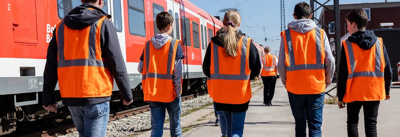 Mehrere Personen in Warnwesten laufen auf dem Werksgelände neben einer S-Bahn