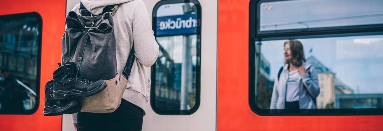 Frau mit Wanderschuhen am Rucksack steht vor der S-Bahn