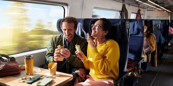 Ein Mann und eine Frau sitzen im ICE und lachen während sie Kleinigkeiten essen