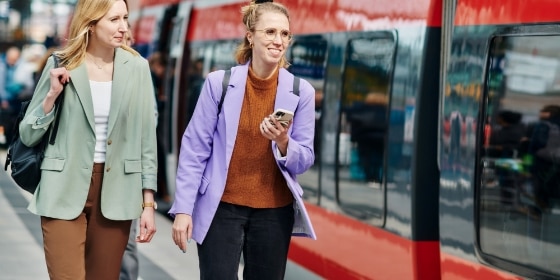 Zwei junge Frauen laufen lachend neben einem Regionalzug am Gleis