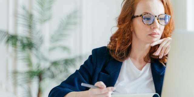 Serious experienced female coach makes records in notepad, focused at screen of laptop computer, has long red hair, wears transparent glasses, watches webinar online. Business and job concept