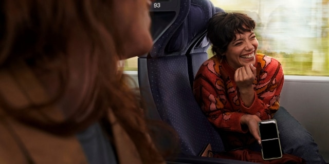 Junge Frau sitzt zum Gang gewendet mit dem Handy in der rechten Hand und lacht andere Passagiere freundlich an